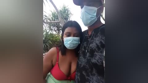 Media: Video of a woman with medium-dark skin and medium build, wearing a red bra and green pants, with a blue mask, next to a man in a black hoodie, standing outdoors in front of palm trees.