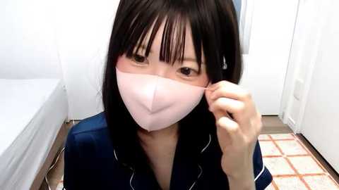 Media: Video of an Asian woman with long black hair, wearing a pink surgical mask, adjusting it with her right hand, indoors, wearing a blue shirt.