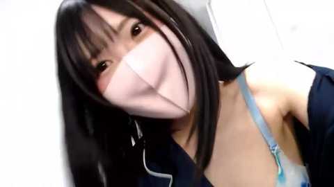 Media: A close-up video of an East Asian woman with straight, dark hair, wearing a light pink surgical mask, blue bra strap, and navy blue jacket.