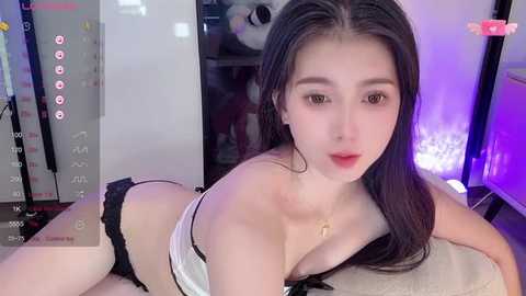 Media: Video of an East Asian woman with long black hair, fair skin, and red lips, wearing a white lace bra and black thong, lying on a beige cushion, in a room with a large panda statue and purple lighting.