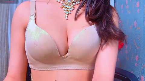 Media: Video of a woman wearing a beige, lacy bra with a deep V-neckline, adorned with a multi-colored necklace. Her skin is light, and her hair is dark and wavy. The background features a textured blue wall.