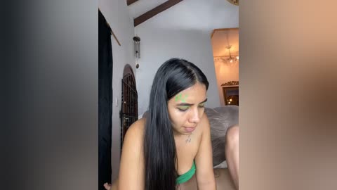 Media: Video of a young woman with long black hair, wearing green lingerie, applying henna to her face in a dimly lit, cozy bedroom with exposed wooden beams and a bed in the background.