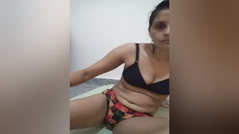 Media: A video of a woman with medium skin tone, black hair, and medium breasts, wearing a black bra and red floral panties, sitting on a bed with white sheets. The background is minimalistic with white walls and a grey carpet.