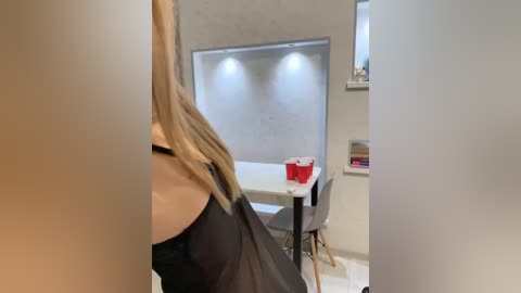 Media: A video of a woman with long blonde hair, wearing a black dress, seated at a small white table in a minimalist, modern room with white walls and a recessed light fixture.