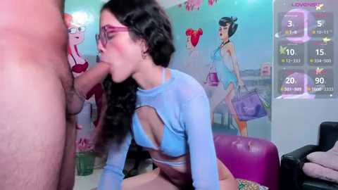 Media: Video of a slender, dark-haired woman in glasses performing oral sex on a nude man in a colorful, cartoon-themed room with a digital game interface.