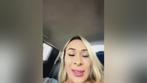 Media: Video of a blonde woman with light skin, closed eyes, and slightly parted lips, captured from a car's rear view mirror. The background shows a blurred interior with a dark car door.