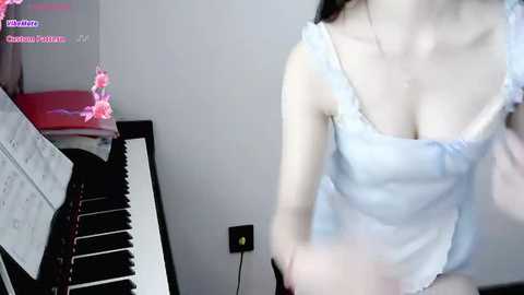 Media: A video of an Asian woman in a light blue, sleeveless top, playing a black upright piano. The room has pink flowers and a pink cushion on the left.