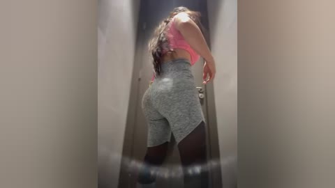 Media: Video of a young woman with long brown hair, wearing a pink crop top and gray high-waisted shorts, walking down a narrow hallway.