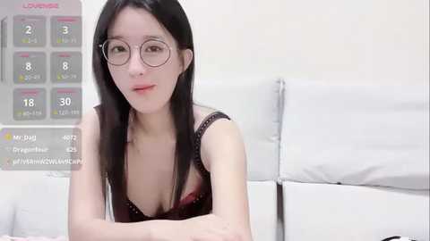 Media: Video of an East Asian woman with long black hair, fair skin, and round glasses, wearing a black lace bra, sitting on a white couch. A calendar with dates and temperatures is visible in the background.