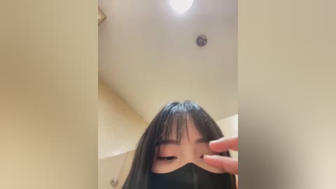 Media: Video of a young Asian woman with long black hair, wearing a black mask, capturing a selfie in a dimly lit room with beige walls and a ceiling light.