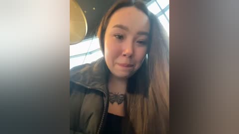 Media: Video of a young woman with straight, shoulder-length brown hair, wearing a green jacket and a black top, with a black lace necklace. She has light skin and is indoors, with a sunlit window and circular lights in the background.