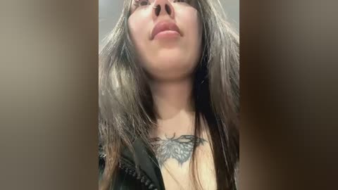 Media: Video of a young woman with long brown hair, wearing a black jacket, showcasing a detailed black butterfly tattoo on her chest. The background is blurry, focusing on her face and tattoo.