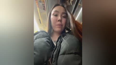 Media: A video of a young woman with long hair, wearing a gray puffer jacket, sitting on a train, with a blurred background.