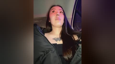 Media: A video of a young Asian woman with long dark hair, wearing a black jacket and a black top, reclining against a dark background. She has a tattoo of a black floral design on her chest.