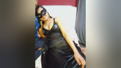 Media: Video of a woman in a black leather catsuit, mask, and high heels, standing in a dimly lit bedroom with red and white bedding.