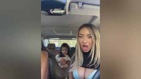 Media: Video of a woman with long blonde hair, large breasts, wearing a low-cut blue top, driving a car. In the background, a young girl with curly hair looks back.