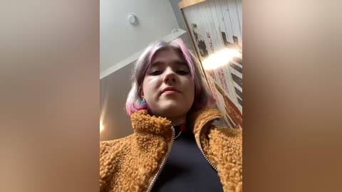 Media: Video of a young woman with pastel pink hair, wearing a fuzzy brown jacket over a black top, standing in a brightly lit room with a colorful wall hanging.