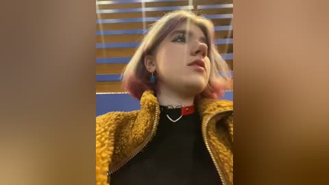 Media: Video of a young woman with shoulder-length, pinkish-blonde hair, wearing a mustard-yellow jacket over a black top, blue earrings, and a red choker. Background features horizontal wooden blinds.