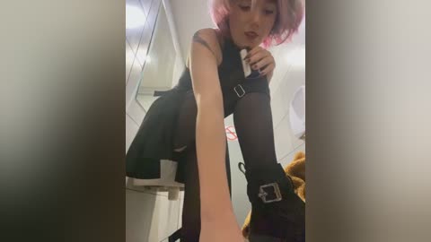 Media: Video of a young woman with short, pink hair and pale skin, wearing a black sleeveless dress, black tights, and black boots with buckles, bending over in a bathroom with a white sink and a teddy bear on the floor.