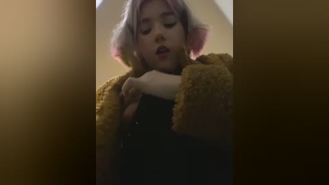 Media: A video of a young woman with pink hair, wearing a fuzzy, mustard-yellow coat, hugging herself in a dimly lit room. Her expression is contemplative.