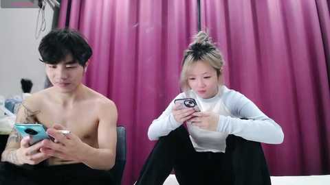 Media: Video of a shirtless East Asian man and a woman with blonde hair, both focused on their phones, seated on black chairs in a room with a pink curtain backdrop.