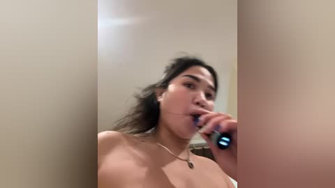 Media: Video of a young, light-skinned woman with dark hair, topless, brushing her teeth in a beige bathroom with a blurred background.