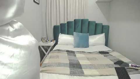 Media: Video of a modern, minimalist bedroom with a teal upholstered headboard, white bedding, and a grey-and-white patchwork quilt. The room features white walls, a small nightstand, and a window with white curtains.