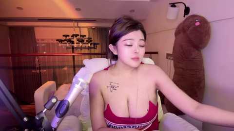 Media: A video of an Asian woman with a fair complexion and medium-length dark hair, wearing a red bra, sitting in a white chair. She has a tattoo on her left shoulder and is being filmed. The background features a modern, dimly lit room with a chandelier.