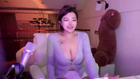 Media: Video of an Asian woman with medium skin tone and dark hair, wearing a low-cut gray cardigan, seated at a desk with a plush bear, illuminated by soft purple lighting.