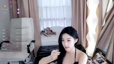 Media: A young East Asian woman with long black hair, wearing a black bra, sits on a bed in a modern, beige-toned room. She touches her hair, with a white chair and curtains in the background.