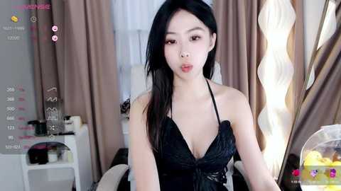 Media: A video of an East Asian woman with long black hair, wearing a black halter dress, in a modern, well-lit bedroom with beige curtains and a white desk.