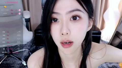 Media: A close-up video of an East Asian woman with long black hair, light skin, and a slender build, wearing minimal makeup. She's indoors, possibly in a bedroom, with a computer monitor displaying social media icons in the background.