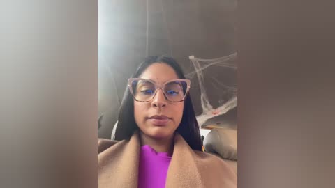 Media: Video of a woman with medium brown skin, long black hair, and large, round, pink-tinted glasses, wearing a beige coat over a bright purple shirt, indoors with a spider web and string lights in the background.