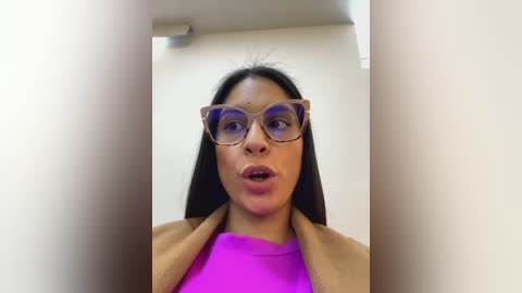 Media: Video of a woman with long black hair, wearing large, light-colored sunglasses and a bright pink shirt, standing in a dimly lit room with white walls.