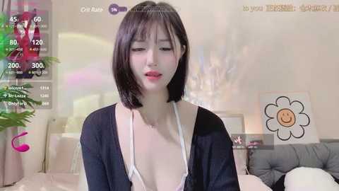 Media: Video of an East Asian woman with short black hair, wearing a white bra and black cardigan, in a cozy bedroom with a floral print pillow and a potted plant.