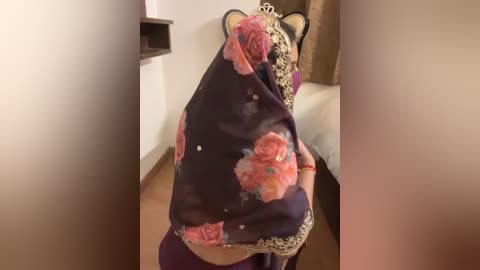 Media: Video of a person wearing a dark purple sari adorned with large, pink floral patterns, standing in a room with beige walls and a wooden floor.