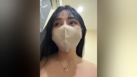 Media: Video of a young Asian woman with long black hair, wearing a beige surgical mask, standing in a bathroom with beige walls, looking up with wide eyes.