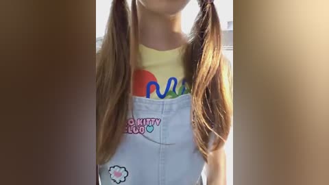 Media: Video of a young girl with long, brown pigtails, wearing a yellow T-shirt with colorful text and white overalls with a pink heart and \"Kitty Love\" patch.