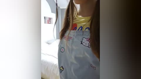 Media: Video of a young girl with long brown hair, wearing a yellow shirt, holding a clear My Little Pony bag. Background features a white bed and light-colored walls.