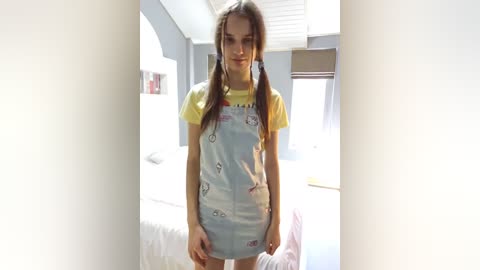 Media: Video of a young woman with long brown hair in pigtails, wearing a yellow T-shirt and light blue overalls with cartoon prints, standing in a bright, modern bedroom with white walls and large windows.