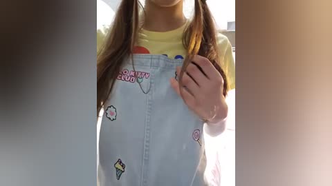 Media: Video of a young woman with long brown hair in pigtails, wearing a yellow t-shirt and light blue overalls adorned with colorful ice cream-themed patches. Sunlight filters through the scene, creating a warm, natural ambiance.