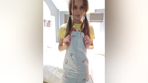 Media: A video of a young girl with long brown pigtails, wearing a yellow t-shirt under a light blue overalls with cartoon designs. She stands in a bright, minimalistic room with white walls and a bed in the background.