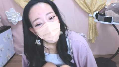 Media: Video of an East Asian woman with long black hair, wearing a light pink robe, white face mask, and pearl earrings. She's seated in a pink room with floral decorations, holding a baby.