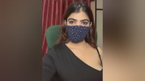 Media: Video of a young woman with light brown skin, long dark hair, and a black one-shoulder top, wearing a blue face mask with white polka dots. Background features a green chair and red curtains.