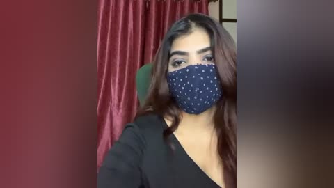 Media: Video of a young South Asian woman with long, wavy brown hair, wearing a navy blue polka-dot face mask, black top, and red velvet curtains in the background.