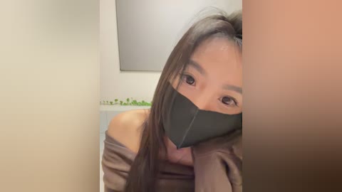 Media: A video of a young Asian woman with long, dark hair, wearing a black face mask and a brown jacket, partially off her shoulders, in a dimly lit room with a white wall and a green plant.