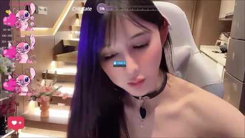 Media: Video of an Asian woman with long black hair and pale skin, wearing a black choker, sitting in a modern kitchen. The background includes white countertops, a refrigerator, and floral decor.