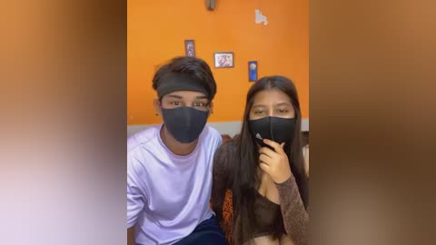 Media: Video of two young people, a boy with short hair in a white shirt, and a girl with long hair in a brown sweater, both wearing black face masks, in an orange-walled room with framed pictures.