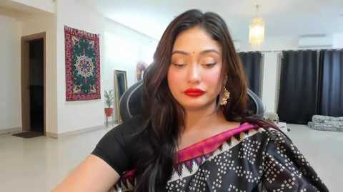 Media: Video of a South Asian woman with medium skin tone, long black hair, wearing a black sari with a maroon border and white geometric patterns, in a modern, minimalist living room with white walls, a chandelier, and a decorative tapestry.