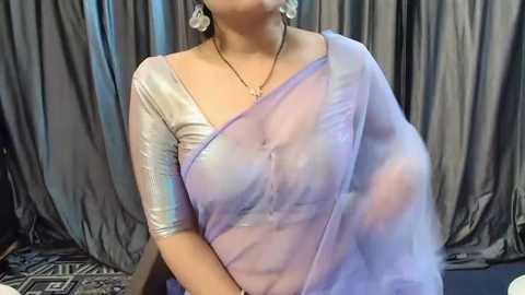 Media: Video of a woman in a sheer lavender sari with a silver blouse, wearing a gold necklace and large earrings, against grey curtains.